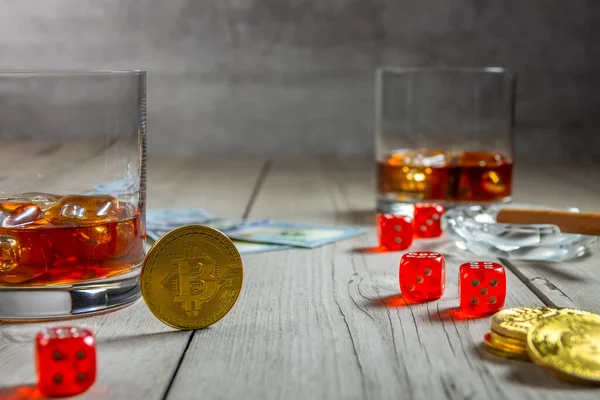 Rustic wooden table. Dice and dollar bills. Two glasses of whiskey with ice cubes and a cigar in an ashtray. Few bitcoin