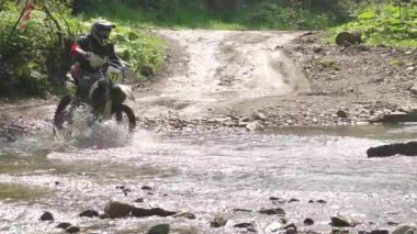 Yaprak döken ormanda güneşli bir yaz günü. Bir enduro motosikletçisi sığ bir nehri bol miktarda su püskürterek geçer. Yavaş çekim