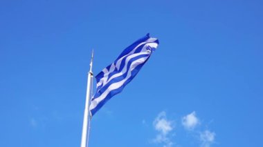 Güneşli bir günde mavi gökyüzü. Bayrak direğindeki Yunan bayrağı rüzgarda dalgalanıyor. Yavaş çekim