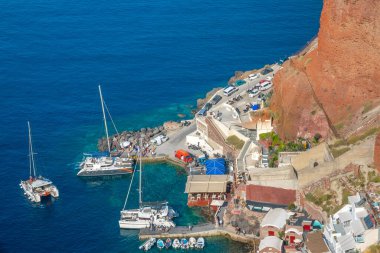 Yunanistan. Santorini 'nin kayalık sahillerinde küçük bir liman. Suda katamaranlar ve motorlu tekneler yüzüyor. Kıyıda bir sürü insan ve araba var.