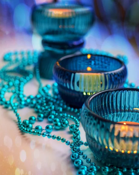 Three cozy lighted candles in turquoise candlesticks with Christmas garland in cold colors with bokeh effect.