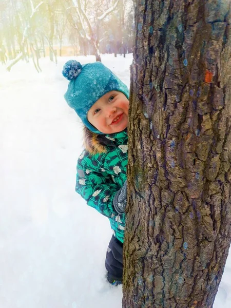 Küçük bir çocuk mutlu bir kış gününde bir ağaç gövdesi arkasından bakmak gülümsüyor