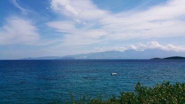 Ön planda beyaz bulutlar ve yeşil bitkiler ile mavi bir gökyüzü altında güzel mavi-turkuaz renk Denizi, Girit