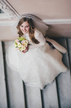 Zarif, zarif bir gelin merdivenlerde aynanın yanında duruyor. Buketi elinde tutuyor. Güzel saç stili ve makyaj. Bir kadın portresi. Kapatın. Portre. Geçmişe dönüş. Klasik mimari içeride..