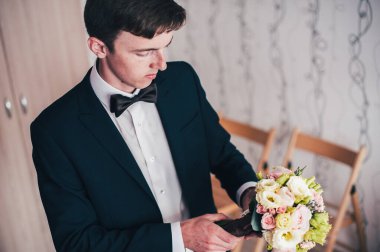 Mutlu damat ayakta duruyor ve poz veriyor. Düğün sabahı. Kapat. Geçmişi. Gülümse, portre. Elinde bir buket gül. Bukete bir bak..