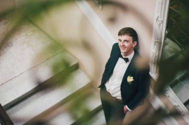 Zarif, zarif bir damat merdivenlerde aynanın yanında duruyor ve parmaklıkları desenli. Cebinde el ele tutuşuyor. Kapatın. Portre. Geçmişe dönüş. Kapalı alanda klasik mimari. Sahte metal merdiven