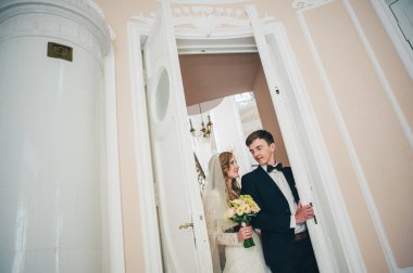 Bir buket çiçekli şık bir gelin damada sarılıyor, ayağa kalkıyor, bakıyor ve kapıyı açıyor. Kapatın. Portre. Geçmişe dönüş. Kapalı alanda klasik mimari. Sütunun yanında.