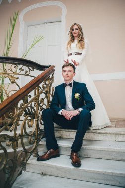 Şık, zarif bir gelin, damadın kafasına boynuzlar takmış elleri merdivenlerde, parmaklıklar ve kapıların yanında desenler var. Kapatın. Portre. Geçmişe dönüş. Kapalı alanda klasik mimari. Sahte metal merdiven