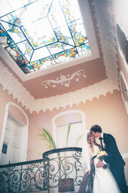 Zarif, zarif bir gelin ve damat merdivenlerde desenli tırabzanlarla dikiliyor. Renkli pencereli bir tavan. Kapatın. Portre. Geçmişe dönüş. Kapalı alanda klasik mimari. Sahte metal merdiven