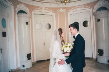 Bir buket düğün çiçeğiyle süslü, zarif bir gelin, çalı gülleri, östoma. Gelin damada sarılıyor. Duvarın arkasında, beyaz kapılar. Kapatın. Portre. Geçmişe dönüş. Klasik mimari içeride.