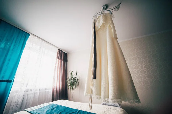 The perfect wedding dress on a hanger on the lamp. Beautiful cream dress with a brown belt and a lace cape. Wedding morning meeting bride. Stylish room in the hotel. With white and blue curtains.