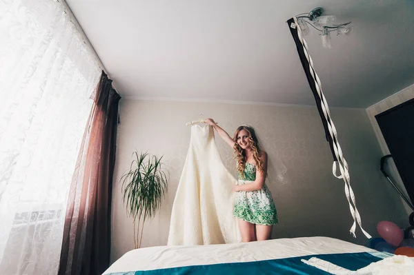 Happy smiling beautiful bride holding a wedding dress on background of white wall. On the veil, hairstyle and makeup. perfect dress on a hanger. lace. Wedding morning meeting bride. Stylish hotel room