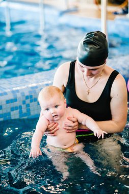 Genç anne, yüzme öğretmeni ve havuzda mutlu küçük bir kız. Çocuğa yüzmeyi öğretiyor. Suda yüzmenin ilk gününün tadını çıkar. Annem çocuğu dalış için hazırlıyor. Egzersiz yapmak