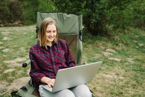 Woman working on laptop online on nature. Young freelancer relaxing in forest. Remote work, outdoor activity in summer. Travel, hiking, technology, tourism, people concept.