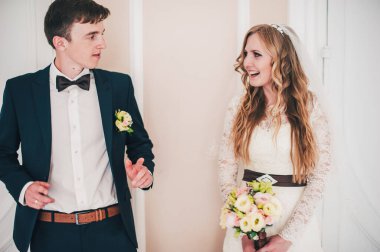 Bir buket düğün çiçeğiyle süslü, zarif bir gelin, çalı gülleri, östoma. Gelin damat gülümsemesi. Duvarın arkasında, beyaz kapılar. Kapatın. Portre. Geçmişe dönüş. Klasik mimari içeride.