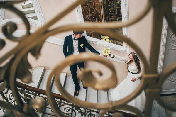 A stylish elegant bride and groom stands near mirror on stairs with railing with patterns. Holds bouquet in hands. hugs. Close up. Portrait. Retro. Vintage architecture indoors. forged metal staircase