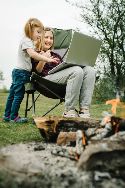 Quarantine, closed nursery school during coronavirus outbreak. Communication with family online on laptop near fire in nature. Homeschooling, freelance job. Mother work on Internet with kid outdoors.