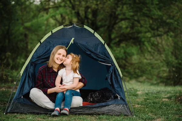 Happy family holidays in a tent with a child in nature. Mother hugs daughter and enjoying a camping holiday in the countryside. Concept of summer vacation and travel, trip. Campsite.