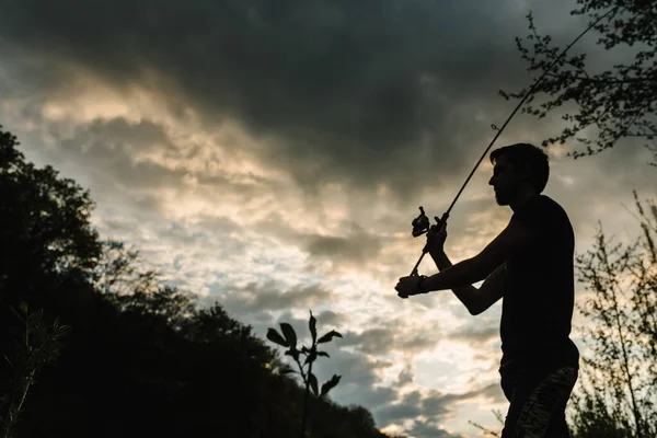 Silhouette of a fisherman. Young man fishing on a lake at sunset. Fishery, fishing day. Rod rings, fishing tackle. Fisherman with rod, spinning reel on the river bank. Fishing for pike, perch, carp.