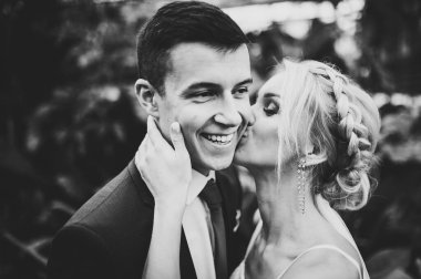 Yeşillik ve yaprakların arka planında duran ve öpüşen damat ve gelinin portresi. Botanik bahçesinde düğün töreni. Siyah beyaz fotoğraf.
