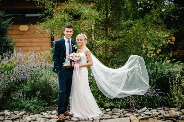 ロマンチックなカップル新婚旅行 花嫁と新郎ピンク 紫色の花や緑 庭のリボンと緑の花束を保持し 結婚式の石の上に立っている — ストック写真