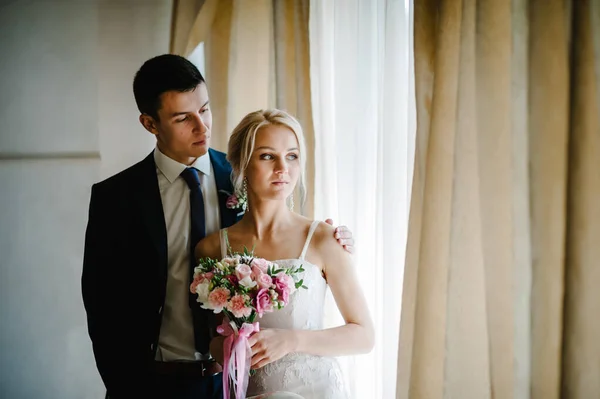 时尚的新娘带着一束花和新郎的婚礼花束靠近房间的窗户 — 图库照片