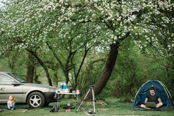 Father working online on the Internet in camp with kid. Quarantine and closed nursery school during coronavirus outbreak. Freelance job. Family vacation with a child near a car and tent.