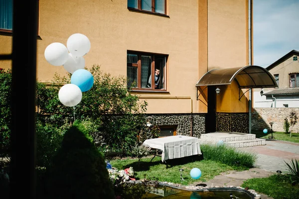Balloons near the fountain in the yard. Fountain made of stones, wood. The man looks from the house. Bride in the window. Wedding ceremony. Wedding Morning, Nature. Close up