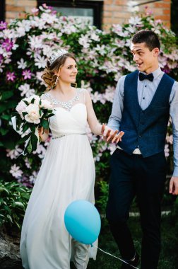 Taçlı ve damatlı şakayık desenli şık bir gelin. Yeni evliler el ele tutuşur. Uçan balon. Kapatın. Clematis. Çiçekler nazikçe pembe ve mordur, yeşil yapraklar.