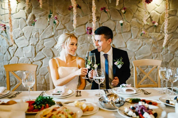 Yeni evliler ellerinde şampanya bardakları tutarak evin içinde kadeh kaldırıyorlar. Çiçekler ve yeşilliklerle süslenmiş düğün töreni. Gelin ve damat restoranın ziyafet salonundaki masada..