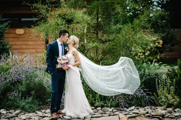 ピンクと紫の花やリボンで緑の花束を保持し 新郎立ってキスを包含し 魅力的な花嫁の肖像画 自然結婚式 — ストック写真