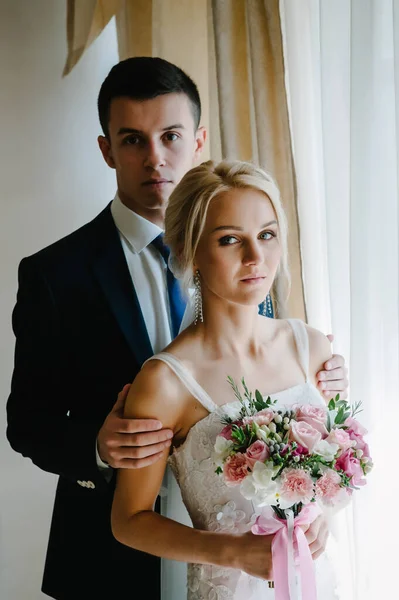 时尚的新娘带着一束花和新郎的婚礼花束靠近房间里的窗户 男人和女人 — 图库照片