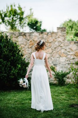 Taçlı şakayık buketli mutlu bir gelin avluda arka planda çimlerin üzerinde dans ediyor. Kapatın. Doğa. Taş duvarın arka planına karşı. Aşağıya bakıyorum..
