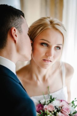 Güzel gelinin ciddi portresi. Bir buket çiçekle ve damat odaların penceresinin yanında. Erkek ve kadın öpüşüyor.