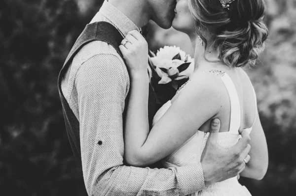牡丹と新郎の花束を持つスタイリッシュな幸せな花嫁 夫は腰の後ろに抱擁し 女性にキス 閉めろ 石垣を背景に 白黒写真 — ストック写真