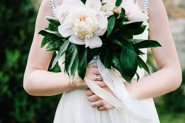 Stylish magnificent bride bouquet of peonies. Close up. On the background of the bride. The image of bride holding a wedding bouquet of pink flowers in hand. A beautiful bouquet. Lace, bow, ribbon