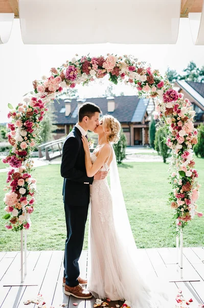 結婚式後の幸せな花嫁と新郎のキス 新婚夫婦だよ 裏庭の宴会場に花や緑で飾られたアーチ — ストック写真