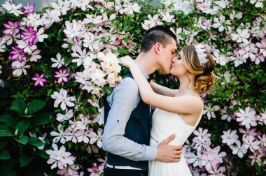 Taçlı ve damatlı şakayık desenli şık bir gelin. Koca kadına sarılır ve onu öper. Kapatın. Clematis. Çiçekler nazikçe pembe, mor ve yeşil yapraklardır. Çalılar, ağaçlar.