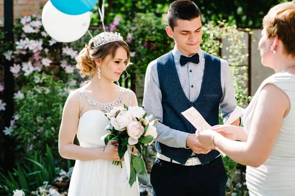 Stylish happy bride with bouquet of peonies with crown, groom hold in hands marriage certificate. woman presented. On background of balloon on nature in courtyard of house. Wedding ceremony. Close up