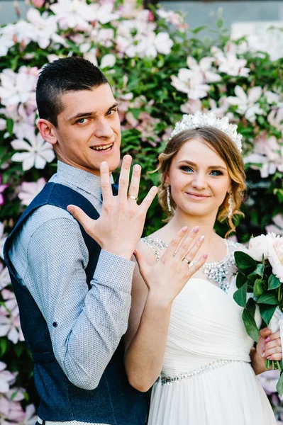Taçlı ve damatlı şakayık desenli şık bir gelin. Yeni evliler altın alyanslarını parmaklarında gösterirler. Kapatın. Clematis. Çiçekler nazikçe pembe ve mordur, yeşil yapraklar.
