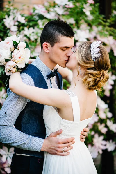 Taçlı ve damatlı şakayık desenli şık bir gelin. Koca kadına sarılır ve onu öper. Kapatın. Clematis. Çiçekler nazikçe pembe, mor ve yeşil yapraklardır. Çalılar, ağaçlar.