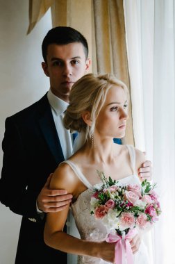 Güzel gelinin ciddi portresi bir buket çiçekle ve damat pencerenin yanında..