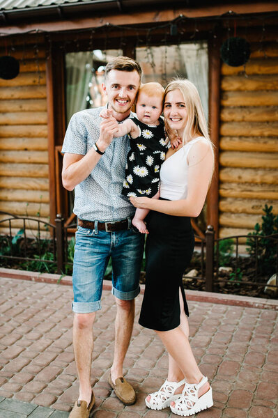 Young beautiful mother and father with a little girl on her hands standing on the background of a wooden house. Baby. Happy family. Full length. Looking the camera. Close up.