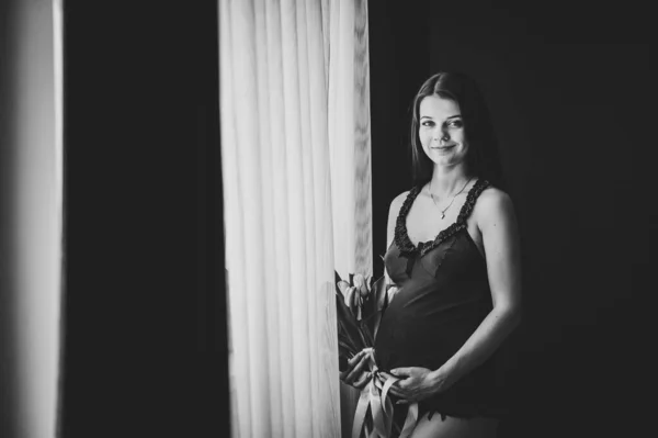 怀孕妇女手里拿着一束鲜花 女孩抱着圆圆的肚子 肚子饿地站在窗前 产妇概念 靠近点9个月 小宝宝洗澡 黑白照片 — 图库照片