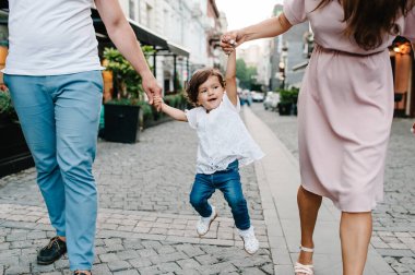 Genç mutlu aile: baba, kız, anne eski şehrin, kasabanın, açık havanın antik mimarisinin yakınındaki bir sokakta yürürken bir bebek fırlatıyor. Aile tatili ve seyahat kavramı.