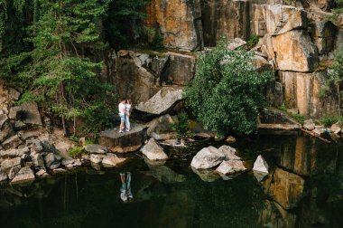 Bir adam ve bir kadın durup kayaların üzerinde, ağacın, ormanın ve gölün yanında sarılıyorlar. Eski bir endüstriyel granit ocağının manzarası. Kanyon. Sonbaharın doğası. Metin ve tasarım için yer. Genç Sevgi Çifti.