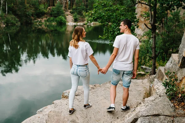 足の若いカップルの石の湖の近くに行く 岩の背景から男と女の視点 古い工業用花崗岩の採石場の風景 閉めろ 人々は手を取り合っていて — ストック写真