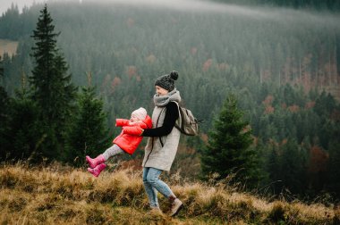 Yüksek kayalıkların sisli zirvesinde sırt çantalı turistler. Mutlu aile: Anne kızını kucağına alır ve çevirir, birlikte vakit geçirir, dağın tepesinde durur. Spor ve aktif yaşam konsepti.