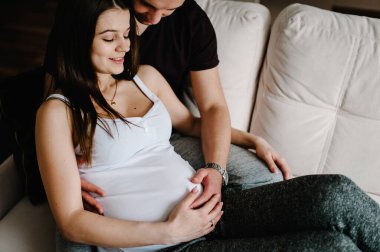 Hamile karısının ellerini karnında tutan bir koca resmi. Hamile kadın ve sevgi dolu yakışıklı adam evde karnına sarılıyor. Sevgi dolu çift. Ebeveynlik kavramı. Bebek Duşu.