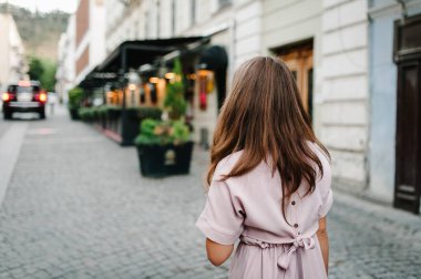 Arka plandaki eski binanın yakınındaki sokakta yürüyen uzun saçlı zarif bir kızın portresi. Şık giyinmiş güzel bir kız..